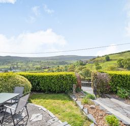 A garden patio with a glass table and chairs overlooking green hills at Henrhiw Bach in Penmachno