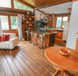 A living area with wooden floors a white couch with cushions a round wooden table and a kitchen with a breakfast bar and stools at Timbertwig Lodge in Lamphey near Pembroke