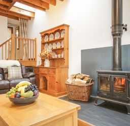 A living room with a wood-burning stove a basket of firewood a leather sofa a wooden cabinet with plates and mugs and a wooden coffee table with a fruit bowl at Pendre Uchaf in Melin-y-Wig near Ruthin