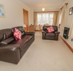 A living room with a leather sofa and armchair with pink cushions and a table by windows at White Rock Cottage in Beadnell