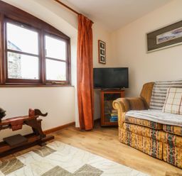 A corner room with a patterned sofa with a pillow and blanket a wooden rocking horse a TV on a wooden cabinet and large window with orange curtain at The Wagon House in Polyphant near Launceston