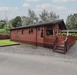 A wooden cabin with a fenced porch and gravel driveway surrounded by trees at Langdale Lodge 15 White Cross Bay near Troutbeck Bridge