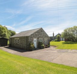 A stone building with a white door and windows next to a wooden shed and a large lawn with trees at Drovers Rest in Otterburn