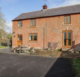 A brick house with wooden windows and doors and a wooden picnic table outside on a paved area at Stables Cottage in Bowerhill near Melksham