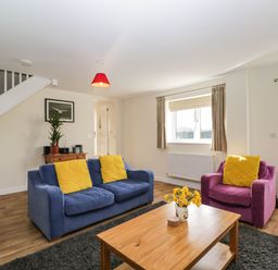 A living room with blue and purple sofas with yellow cushions a wooden coffee table with flowers and a staircase at Stables Cottage in Bowerhill near Melksham