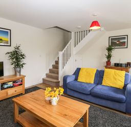 A living room with a blue sofa and yellow cushions a wooden coffee table with flowers television on wooden stand and stairs at Stables Cottage in Bowerhill near Melksham