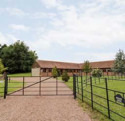 A gravel driveway with a black metal gate leading to a single-story brick and stone building with a pitched roof surrounded by grass and trees at Beesoni Lodge in Castlemorton near Malvern
