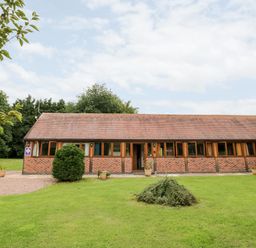A single-story brick building with multiple windows and a tiled roof surrounded by grass and trees at Beesoni Lodge in Castlemorton near Malvern