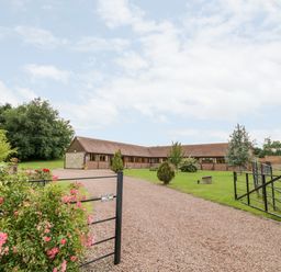 A gravel driveway with a black metal gate leading to a single-story building with a tiled roof and brick walls at Beesoni Lodge in Castlemorton near Malvern