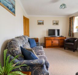 A living room with a patterned sofa, a television on a wooden cabinet, a leather chair, and a window with patterned curtains at 1 Cowdea Farm in Bettiscombe near Beaminster and Bridport