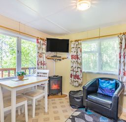 A small dining table with two chairs beside windows with floral curtains a wall-mounted television an electric fireplace a black armchair with a blue cushion and a window at Bluebell Cottage in Maenan near Llanrwst