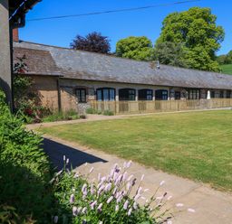 A long brick and flint building with multiple doors and windows a grass lawn and flowers at The Old Stables in Winterborne Stickland