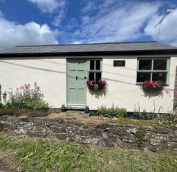 A small building with a green door and two windows with flower boxes a stone wall and garden in front at Palmers Lodge in Egloskerry near Launceston