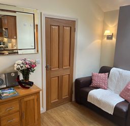 A small living room with a wooden door a brown leather sofa with pillows a wooden cabinet with flowers and a mirror above at Palmers Lodge in Egloskerry near Launceston