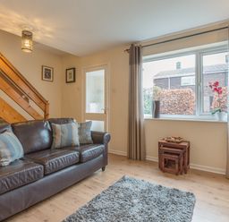 A living room with a brown leather sofa and pillows a gray rug wooden stairs and a window with curtains at Crumstone Corner in Seahouses