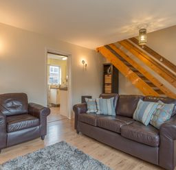 A living room with a brown leather sofa and armchair near wooden stairs and an open door to a kitchen at Crumstone Corner in Seahouses