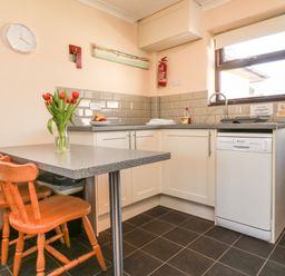 A kitchen with a table and two wooden chairs a vase with red flowers on the table a countertop with a sink dishwasher and a window at Honeysuckle Cottage in Bude