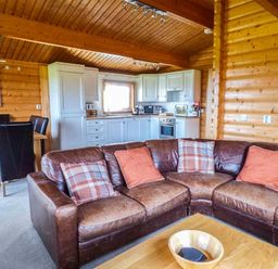 A wooden interior with a brown leather corner sofa with cushions a wooden dining table with black chairs and a white kitchen at Ty Pren Llanrhian near St Davids
