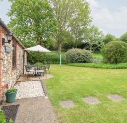 A garden with a stone and brick cottage patio table chairs and umbrella and green lawn at Moonlight Cottage in Coltishall