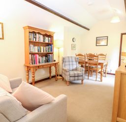 A living room with a sofa a plaid armchair a wooden bookshelf and a dining table with chairs at Moonlight Cottage in Coltishall