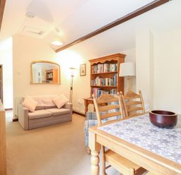 A living room with a beige sofa a wooden bookshelf a dining table with chairs and a bowl on top at Moonlight Cottage in Coltishall