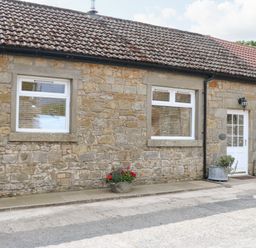 Stone cottage exterior with two windows and a white door with flower pots at Stable Cottage in Belford near Bamburgh