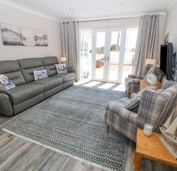 A living room with a leather sofa, two plaid armchairs, a wooden side table with a model sailboat and candles, a wall-mounted TV, and French doors with curtains at St Winifreds in Rhosneigr