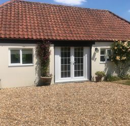 A single story building with a tiled roof two windows and a double glass door with plants and flowers outside at Oke Apple Cottage in Okeford Fitzpaine