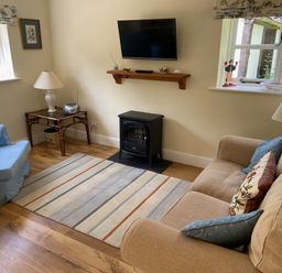 A living room with a beige sofa a blue armchair a striped rug a wall-mounted TV and a small stove at Oke Apple Cottage in Okeford Fitzpaine