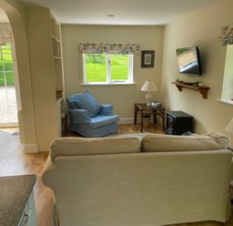 A living room with armchair sofa lamps television and windows showing grass at Oke Apple Cottage in Okeford Fitzpaine