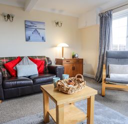 A living room with a black leather sofa with cushions a wooden coffee table with a basket and mugs a grey armchair next to a window a wooden sideboard with a lamp and decor at Glan Y Don Bach in Abersoch