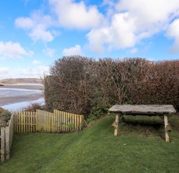 A garden area with a wooden picnic table and wooden fence overlooking a beach and water at Glan Y Don Bach in Abersoch