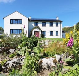 A white two-story house with several windows and a front door surrounded by greenery and colorful flowers at Ghyll Bank House in Staveley near Windermere