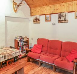 A room with a red three-seater sofa and pillows a small bookshelf filled with books a table with a tablecloth and placemats wooden ceiling and framed pictures on the wall at Willowbrook Cottage in Askill County Leitrim
