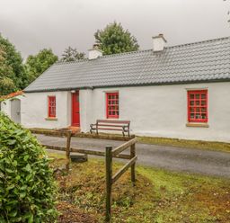 A white cottage with a gray roof red door and red window frames a bench outside the house on a gravel driveway with greenery around at Willowbrook Cottage in Askill County Leitrim