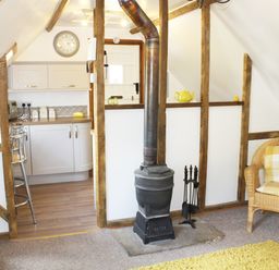 A room with a wood stove a wicker chair and a kitchen area at The Hayloft in Necton near Swaffham