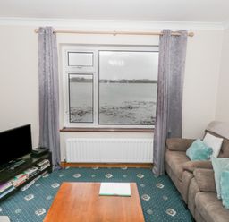 A living room with a sofa a wooden coffee table a television on a stand and a large window overlooking water at Quayside House in Roundstone County Galway
