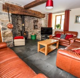 A living room with leather sofas a wooden coffee table a stone fireplace with a wood burner a television and red curtains at Maestyddyn Farm House Clawddnewydd near Ruthin