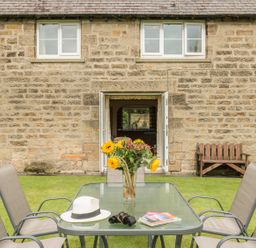 A garden table with chairs a vase of sunflowers a white hat binoculars and magazines outside a stone house with open doors at Brian's Cottage in Alnmouth