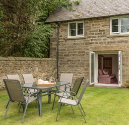 A patio table with six chairs on grass outside a stone house with open doors showing a sofa inside at Brians Cottage in Alnmouth