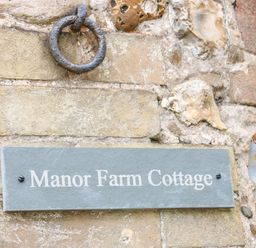 A stone wall with a metal ring above a sign that reads Manor Farm Cottage in Swaffham
