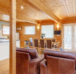 A wooden cabin interior with a dining table and chairs a kitchen area with appliances and two brown leather chairs at Jamaal Lodge in South Broomhill near Amble