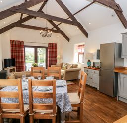 A dining table with six chairs set with plates and glasses in an open plan kitchen and living area with wooden beams on the ceiling at Potters Barn in Begelly near Kilgetty
