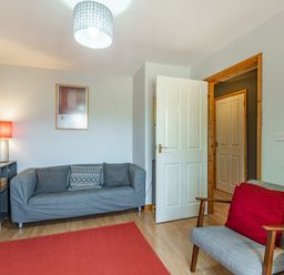 A sitting room with a grey sofa, an armchair with a red cushion, a red rug, a bookshelf, and a standing lamp at Gorwel in Llan Ffestiniog near Blaenau Ffestiniog