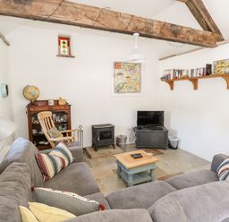 A living room with a large gray sectional sofa, wooden rocking chair, small coffee table, wood stove, TV on a stand, bookshelf, and exposed wooden beams at The Visiting House in Gorteen near Dunmore County Galway