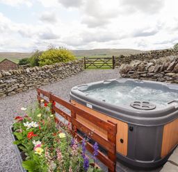 An outdoor hot tub next to a flower bed with a stone wall and green fields in the background at Y Bwthyn in Ysbyty Ifan