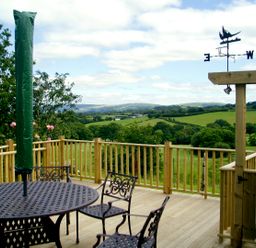 A wooden deck with metal table and chairs and a weather vane overlooking green fields and hills at Cwm Derw Cottage Llanafan Fawr near Builth Wells