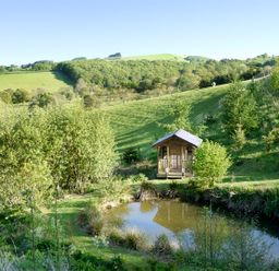 A small wooden cabin near a pond with trees and grassy hills at Cwm Derw Cottage Llanafan Fawr near Builth Wells