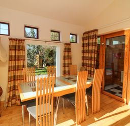 A dining room with a glass table and wooden chairs next to windows and wooden doors at Sutton Cottage Ballysheen near Rosslare Harbour County Wexford