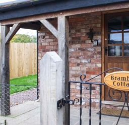 The entrance of a brick cottage with a wooden door and a metal gate with a sign reading Brambles Cottage at Little Malvern near Malvern
